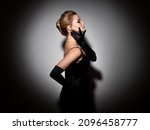 Small photo of A femme fatale in a black dress with an open back and long velvet gloves. Attractive young blonde with makeup for a celebration, portrait in a spot of light