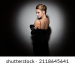 Small photo of A femme fatale in a black dress with an open back and long velvet gloves. Attractive young blonde with makeup for a celebration, portrait in a spot of light