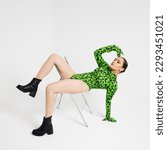 Small photo of Stylish bright dancer in a bright green leopard print bodysuit, posing on a chair, studio photo on a white background. Plastic body, pretentious pose