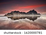 Panorama of Famous Stokksnes mountain reflected in water on Vestrahorn cape (Batman Mountain), Iceland, Europe. Popular tourist attraction. Beauty world. Red sky over black sand dunes on the beach. 