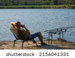 Small photo of Fisherman with hot drink in mug waiting a bite with carp fishing equipment. Angler is fishing with carpfishing technique in a beautiful summer evening, using rod pod, bite alarms, swinger, rods
