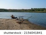 Small photo of Fisherman with hot drink in mug waiting a bite with carp fishing equipment. Angler is fishing with carpfishing technique in summer evening, using rod pod, bite alarms, swinger, rods, landing net