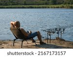 Small photo of Angler with hot drink in mug waiting a bite with carp fishing equipment. Angler is fishing with carpfishing technique in a beautiful summer evening, using rod pod, bite alarms, swinger, rods