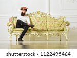 Small photo of Macho sits with open book on head, like roof. Man with beard and mustache sits on sofa, white wall background. Overstudy concept. Guy overdid with studying, fed up of reading old boring book.
