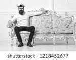 Small photo of Macho sits with open book on head, like roof. Overstudy concept. Guy overdid with studying, fed up of reading old boring book. Man with beard and mustache sits on sofa, white wall background.