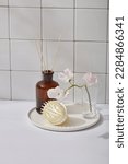 Small photo of Reed air freshener in amber bottle unlabeled, white scalp massage brush and beauty flower branch on round dishes, decorated on white tile wall background. Aromatherapy concept to freshen the room