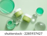Two unlabeled jar placed on white background with some glass balls, green liquid contained in petri dishes and erlenmeyer flask. Mockup of skin care or body care product