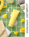 Small photo of Unbranded plastic tube decorated with toy yellow duck, towel and hair tier, mirror and scalp massage brush on green ceramic background. Mockup of skin care cosmetic tube of beauty facial