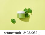 View from above of an unbranded moisturizer jar with fresh pennywort leaves standing out against a pastel background. Branding with blank labels.
