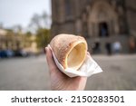 Man holding a touristic trdelnik spit cake with catholic Church of St. Ludmila in the background, Prague, Czechia