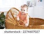 Small photo of a cute little girl child in a pink bodysuit is standing in a wicker cradle at home, looking at the camera, smiling, children's healthy sleep