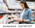 Portrait young woman in supermarket holding two bottles of yogurt reads labels compares chooses healthy dairy food