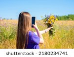a teenager girl or young woman with beautiful long hair takes a photo of a bouquet of wild flowers on her mobile phone. creates content for his channel, walks in a flowering field.
