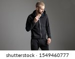 young casual man standing and fixing his tracksuit while looking aside pensive on gray studio background