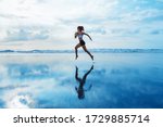 Small photo of Barefoot young girl with slim body running along sea surf by water pool to keep fit and burning fat. Beach background with blue sky. Woman fitness, jogging sports activity on summer family vacation.