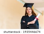 Smiling female Asian student in academic gown and graduation cap holding diploma