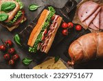 Delicious baguette sandwich with ham, bacon, cheese, lettuce, tomatoes, sausage, gammon on cutting board with herb and spices over on dark background. Meat food, top view, flat lay, toning
