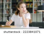 School girl wearing white t-shirt sitting at table and writing in exercise book, looking to side and daydreaming, bookshelves on blurred background. Pupil doing assignment in class