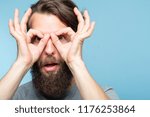 Small photo of funny ludicrous joyful comic playful man pretending to look through binoculars made of hands. portrait of a young bearded guy on blue background. emotion facial expression concept
