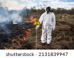 Small photo of Firefighter ecologist extinguishing fire in field. Man in protective radiation suit and gas mask near burning grass with smoke, holding warning sign with exclamation mark. Natural disaster concept.