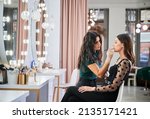 Small photo of Makeup artist doing professional makeup in visage studio. Stylish woman sitting at dressing table while female beauty specialist in sterile gloves applying lipstick on client lips with cosmetic brush.
