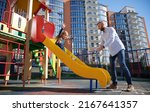 Small photo of Handsome dad catching cheerful daughter, riding on slide at modern courtyard of city residential high-rise buildings. Bearded father catching pretty girl while standing near slide on playground.