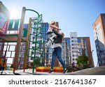 Small photo of Happy blond mom catching cheerful daughter, jumping from ladder at playground. Beautiful woman having fun with female child at modern courtyard of city residential high-rise buildings.