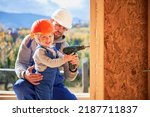 Small photo of Father with toddler son building wooden frame house. Male builder and kid using screwdriver on construction site, wearing helmet and blue overalls on sunny day. Carpentry and family concept.