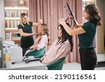 Small photo of Female hairstylist curling client hair with curling iron device while colleague combing woman hair. Professional hairdresser doing hairstyle for woman in beauty salon.
