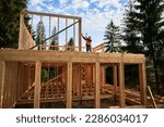 Wooden two-story, frame-built home is being erected by the carpenter near the forest. Man is driving in nails with hammer. Underpinning idea of this venture is modern, ecological building practices.