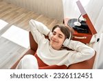 Handsome teenager boy wearing earphones listening vinyl records on retro wooden turntable, laying on a brown sofa and smiling, enjoy music sound