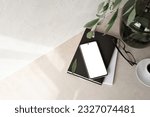 Mobile phone with empty screen mockup, notebook, glasses, vase with foliage branch, empty neutral beige background with aesthetic sunlight shadow. Minimalist business brand, social media blog template