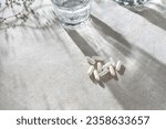 White capsules, glass with water and dried grass on neutral beige linen tablecloth with aesthetic light shadows.