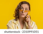 Small photo of Tired sleepy young woman in glasses talking on mobile phone with friend making online conversation. Disinterested attractive girl having annoyed boring talk on smartphone isolated on yellow background