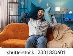 Small photo of Repair work at neighbours. Irritated young man relaxing on couch cover ears with pillows annoyed by noisy neighbors suffer from headache wish silence. Thin walls at home flat without sound insulation