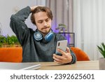 Small photo of Sad displeased Caucasian man use smartphone typing browsing, loses becoming surprised sudden lottery game results, bad news, fortune loss fail, deadline, virus. Young adult guy at home office at table