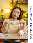 Small photo of Poor young woman insufficient amount of money, holding piggybank and one dollar banknote. Financial crisis. Bankruptcy. Poverty and destitution. Female girl sitting at home office at table. Vertical