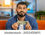 Small photo of Portrait of poor Indian Arabian man upset over insufficient amount of money showing one dollar banknote. Financial crisis. Bankruptcy. Poverty destitution. Upset Hispanic guy sitting on sofa at home