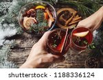 first person view of mans and womans hands holding glasses with Christmas hot mulled red wine with spices and fruits on a wooden rustic table background with spruce snow-covered branches. Traditional