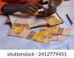 Small photo of Black person in orange long sleeves shirt working on desk, holding mobile phone and touching on screen with Ugandan shilling notes on desk. accounting concept