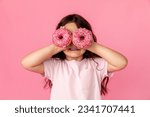 Portrait of a little smiling girl with curly hair and two appetizing donuts in her hands, closes her eyes with donuts, on a pink background, a place for text. Dieting concept and junk food.