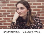 Small photo of Close up portrait of joyless 20s woman sitting alone near indoor wall. Face of caucasian serious female with sad eyes staring away from camera. Tiredness, lack of optimism, solitude, life concerns
