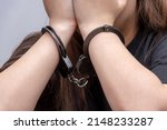Small photo of A young girl handcuffed hides her face on a gray background, close-up. Juvenile delinquent in a black T-shirt, criminal liability of minors.