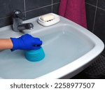 Small photo of White sink with water and blue plunger in hand clears blockage. Plumbing tool for mechanical cleaning of blockages in sewer pipes and removal of mud from them, preventing the movement of water.