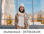 stylish attractive young smiling woman walking in street in winter outfit with coffee wearing checkered coat, white knitted hat and scarf, happy mood, fashion style trend