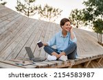 young pretty woman working on laptop in IT outside in modern park street sitting on remote work, summer style casual smiling talking on wireless earpods drinking coffee