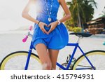 young beautiful hipster woman posing with bicycle on beach, showing heart with hands, gesture, concept, details, summer vacation, vintage style, boho outfit, blue dress, slim body, bright, colorful
