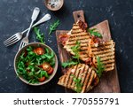 Grilled bacon, mozzarella sandwiches on wooden cutting boards and arugula, cherry tomato salad on dark background, top view.Delicious breakfast or snack, flat lay    