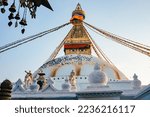 View of the Boudhanath Stupa in Kathmandu Nepal against a blue sky in warm evening light. There are many pigeons on the roof. In the foreground blurred bells.