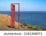 An old red open door stands on a cliff near the blue sea